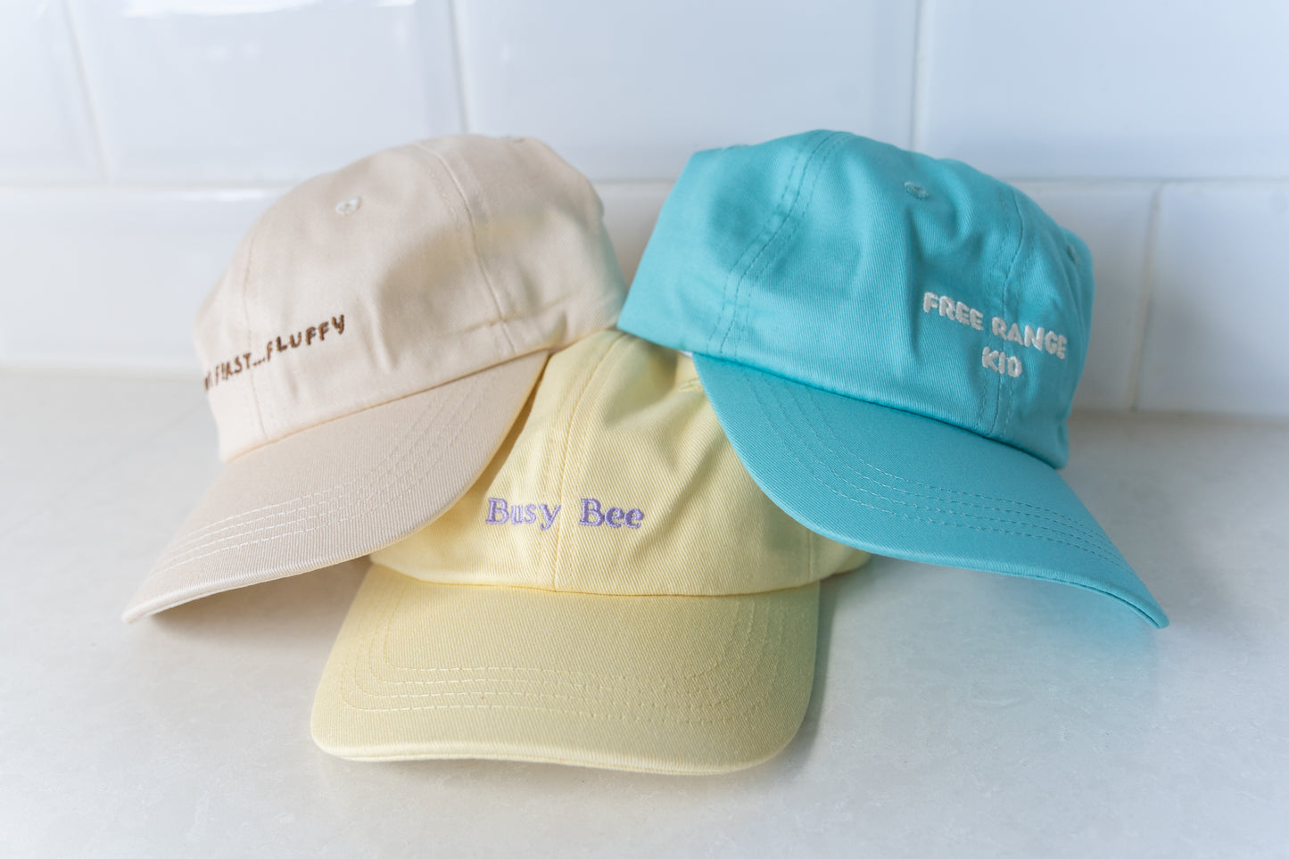 Three kids' hats featuring 'But First, Fluffy,' 'Free Range Kid,' and 'Busy Bee' slogans, perfect for playful and adventurous children.