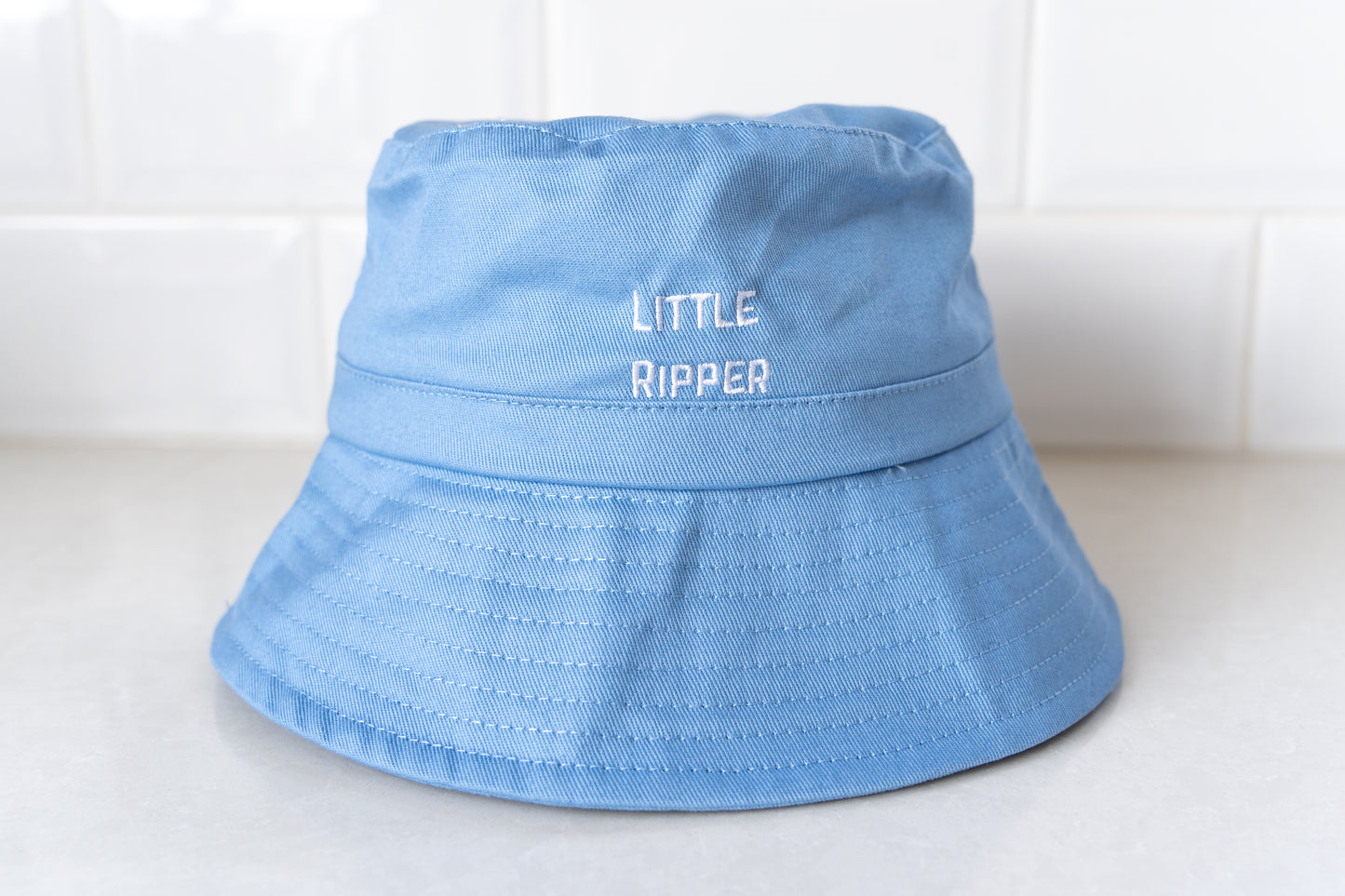 Front view of blue 'Little Ripper' bucket hat for kids with white embroidered text, designed for sun protection and outdoor play.